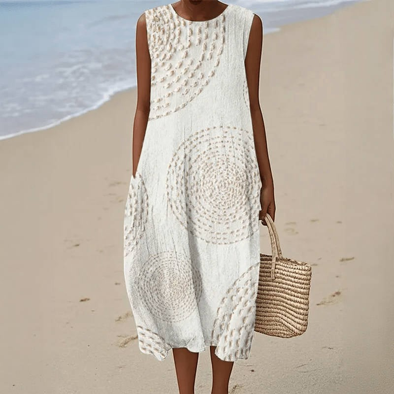 Victoria Boutique sleeveless midi dress in white with a beige circular print, featuring side pockets on a beach.